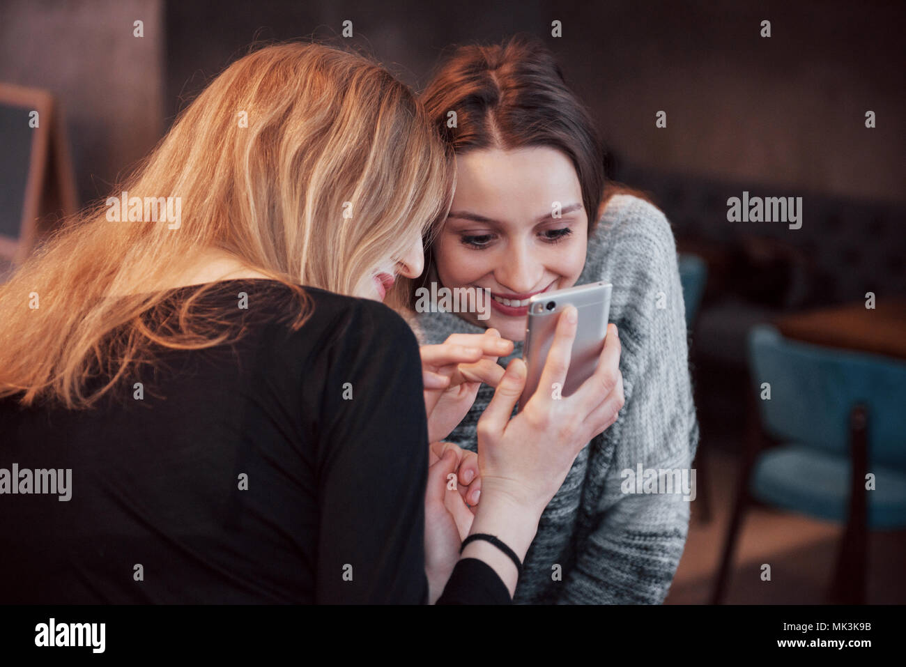 Dos sonriendo amigos leer funny chat sobre teléfono moderno sentado con sabroso café en el restaurante.Hipster niñas disfrutando de tiempo de recreo en la cafetería con bebidas calientes y mensajería en teléfonos Foto de stock