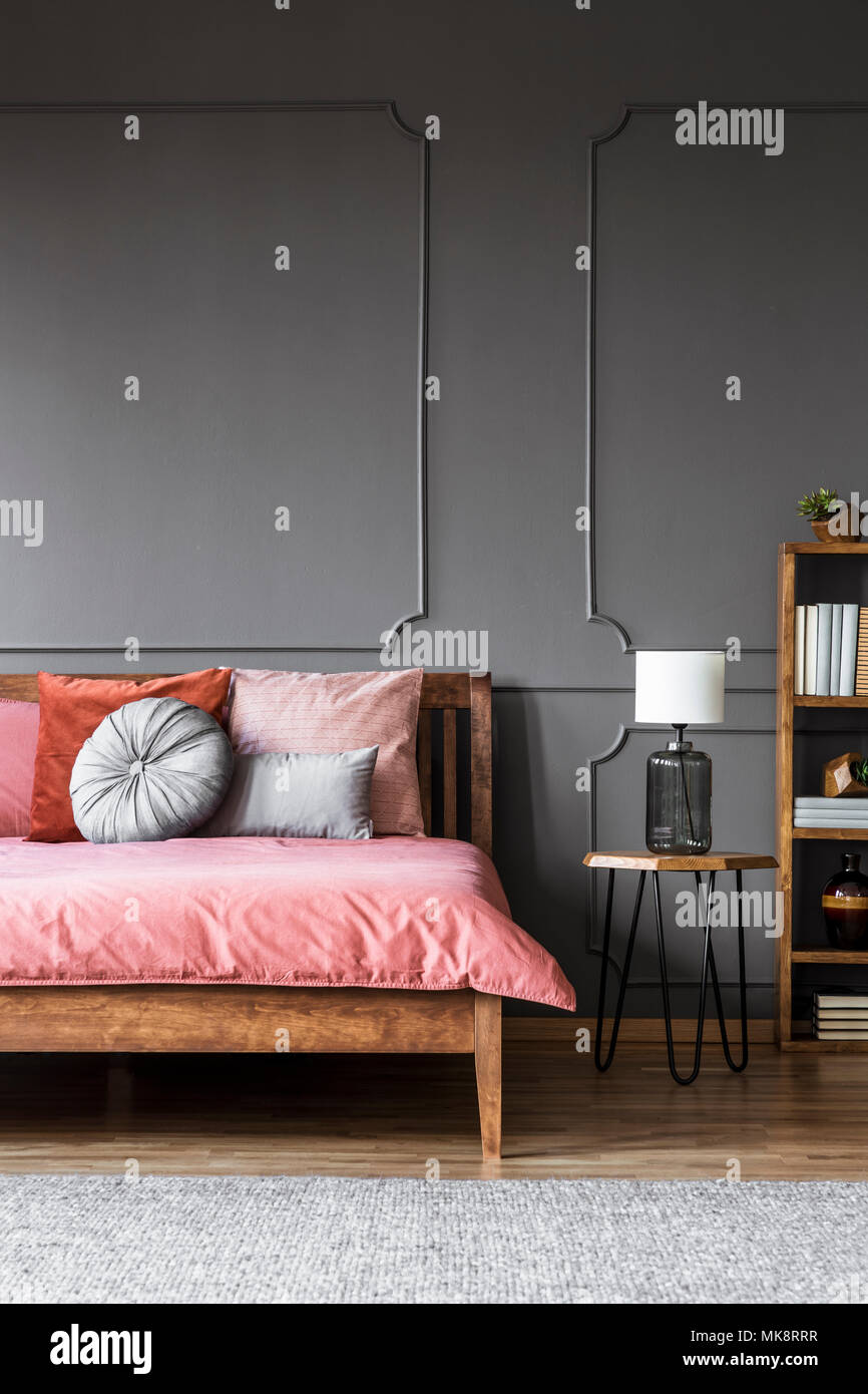 Lámpara blanca sobre un taburete junto a una cama de madera con sábanas de color rosa en gris oscuro interior del dormitorio con el moldeado en la pared Foto de stock