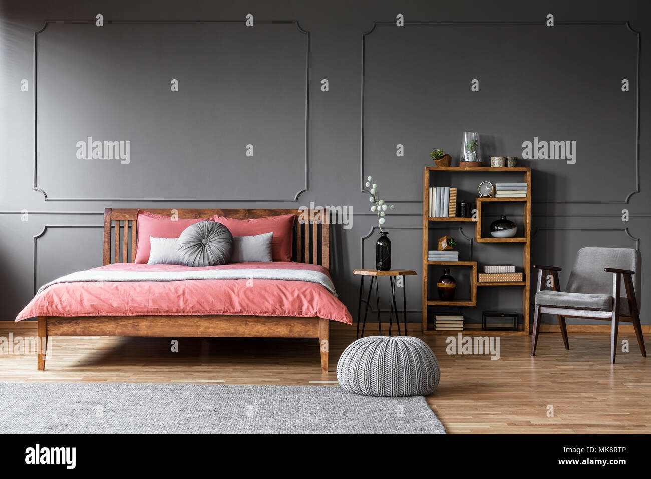 Puf gris cerca de la cama de madera con sábanas de color rosa en el elegante interior del dormitorio con sillón Foto de stock