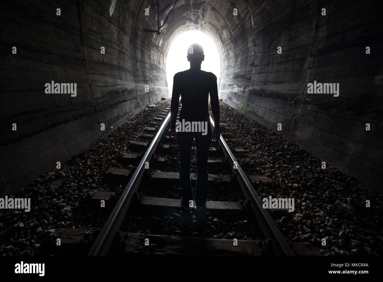 Hombre silueteado en un túnel de pie en el centro de la vía férrea hacia la luz al final del túnel en una imagen conceptual Foto de stock