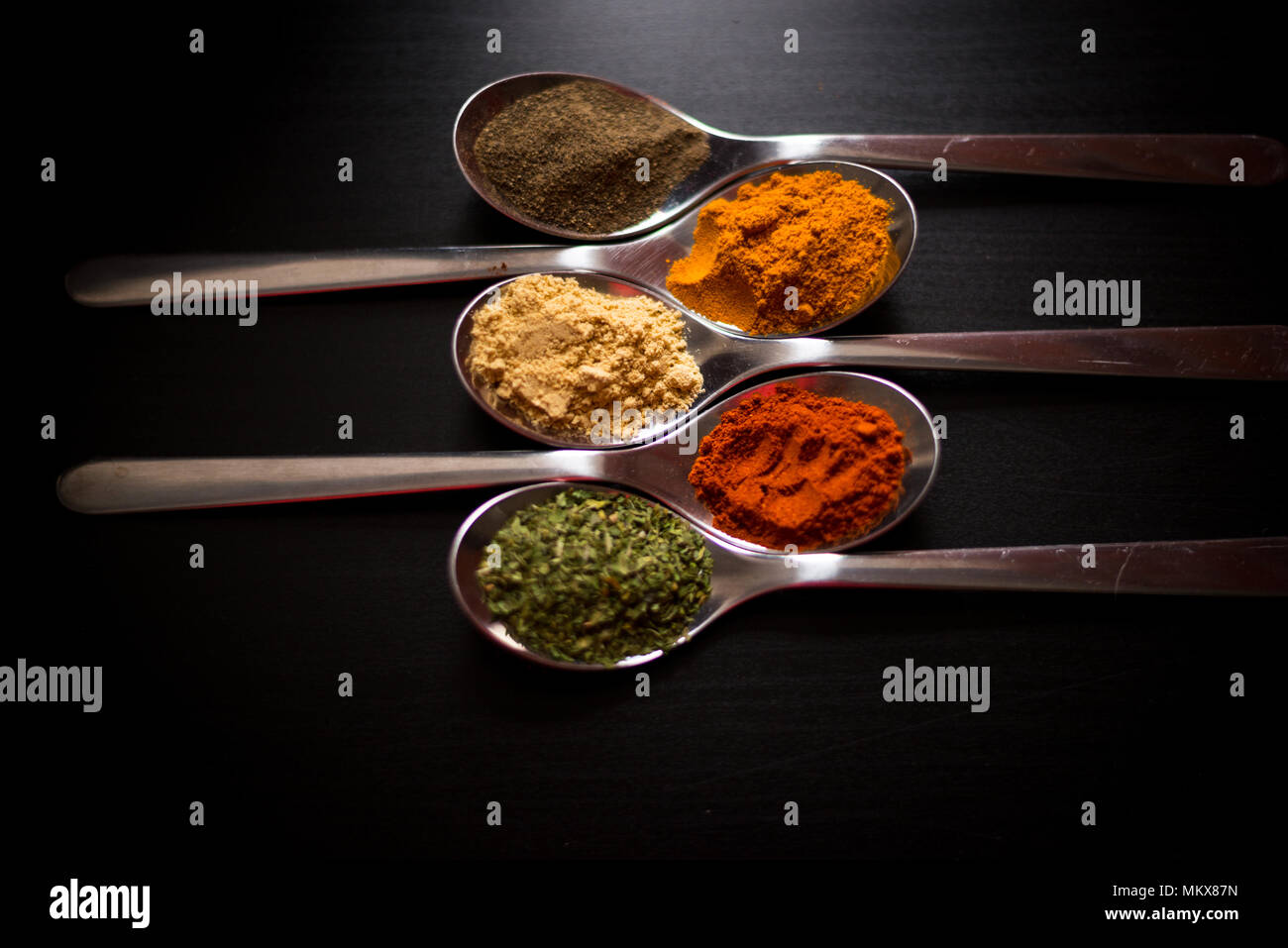 Especias coloreadas en cucharas de plata, colocados sobre una mesa de madera negra. Pimienta, pimienta, sal, albahaca, mostaza Foto de stock