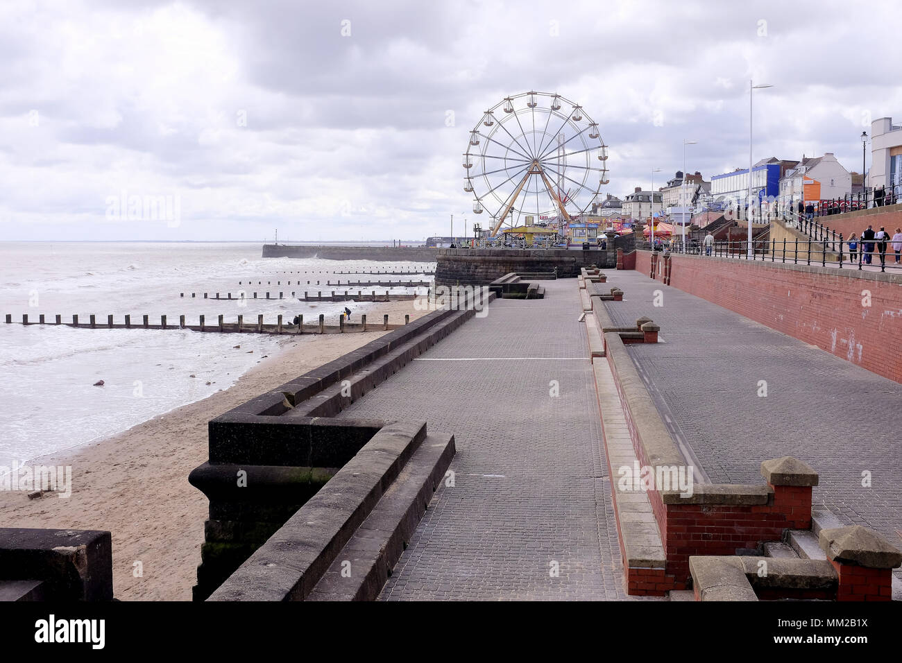 Bridlington, Yorkshire, Reino Unido. El 29 de abril de 2018. Los turistas caminando por el paseo marítimo y el parque de atracciones big wheel siluetas contra un cielo nublado . Foto de stock