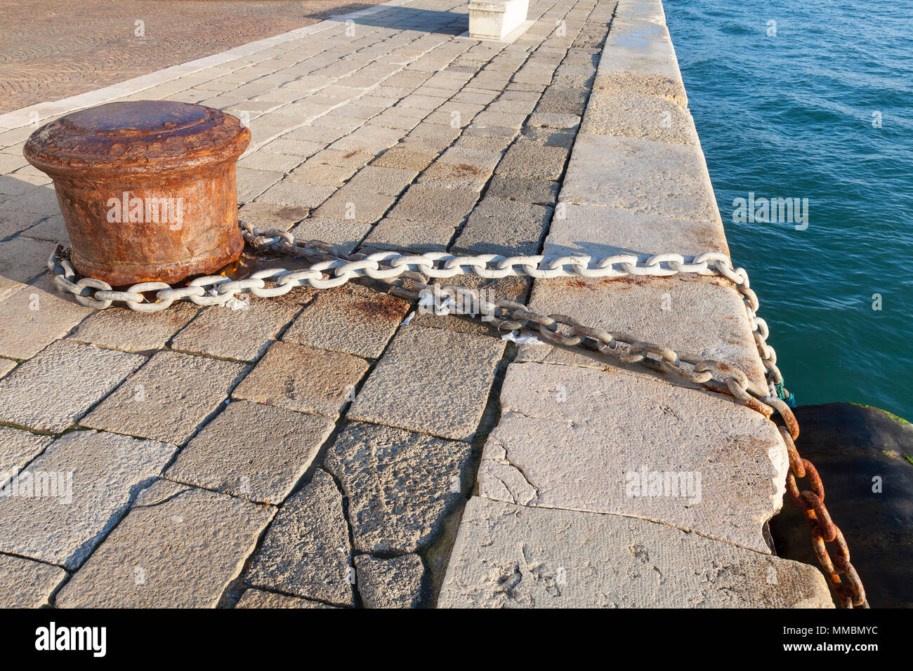 El hierro viejo bolardo con cadena de eslabones metálicos conectados y grandes guardabarros, causando daño a la piedra de Istria quay en el borde de la laguna veneciana, Castell Foto de stock