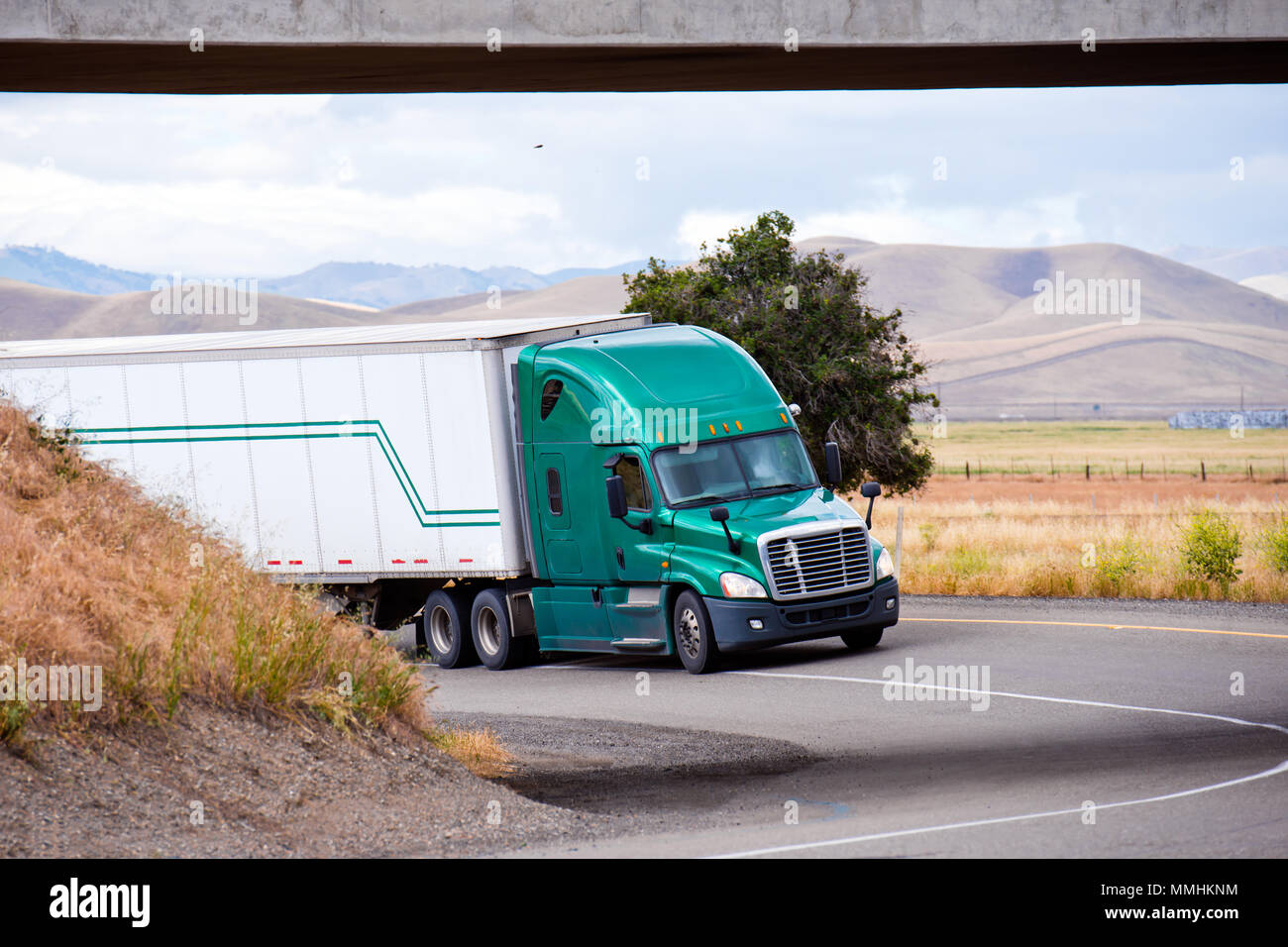 Big Rig verde larga semi camión con semirremolque furgón seco de transportar la carga comercial en día soleado en California y pasando por debajo del puente de la ACR Foto de stock