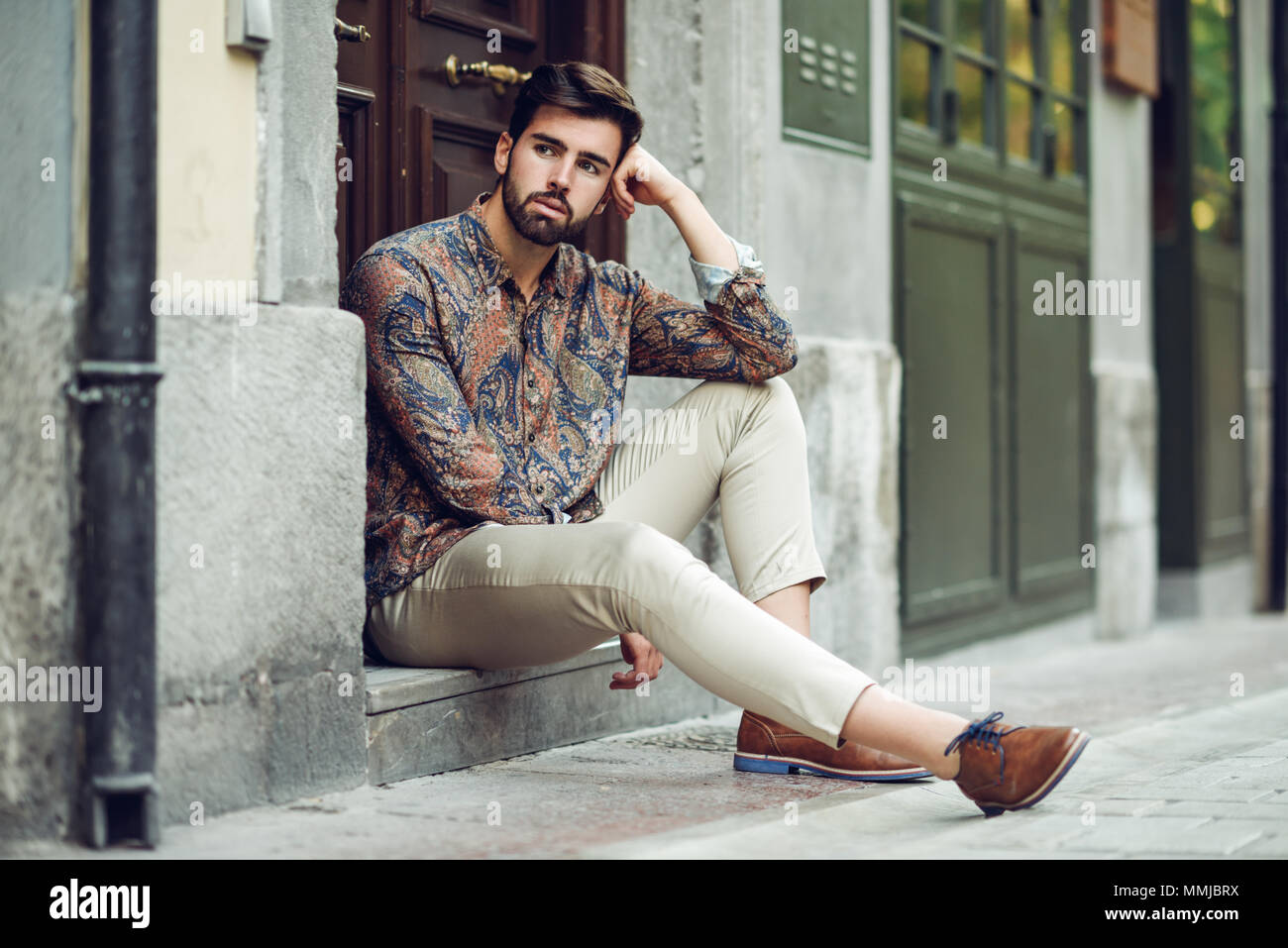 Joven Hombre Barbado, modelo de moda urbana, sentados en un paso vistiendo ropa casual. Chico con barba y peinado moderno mirando lado de la calle Fotografía de stock - Alamy