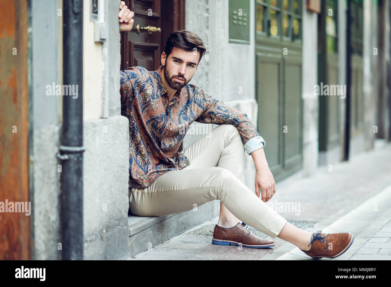 Joven Hombre Barbado, modelo de moda urbana, sentados en un paso vistiendo ropa  casual. Chico con barba y peinado moderno mirando a otro lado de la calle  Fotografía de stock - Alamy