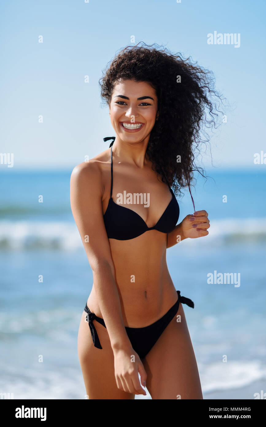 Joven mujer árabe con hermoso cuerpo en traje baño sonriendo en una playa tropical. Morena hembra con rizado peinado largo vistiendo negro bikini Fotografía de stock -