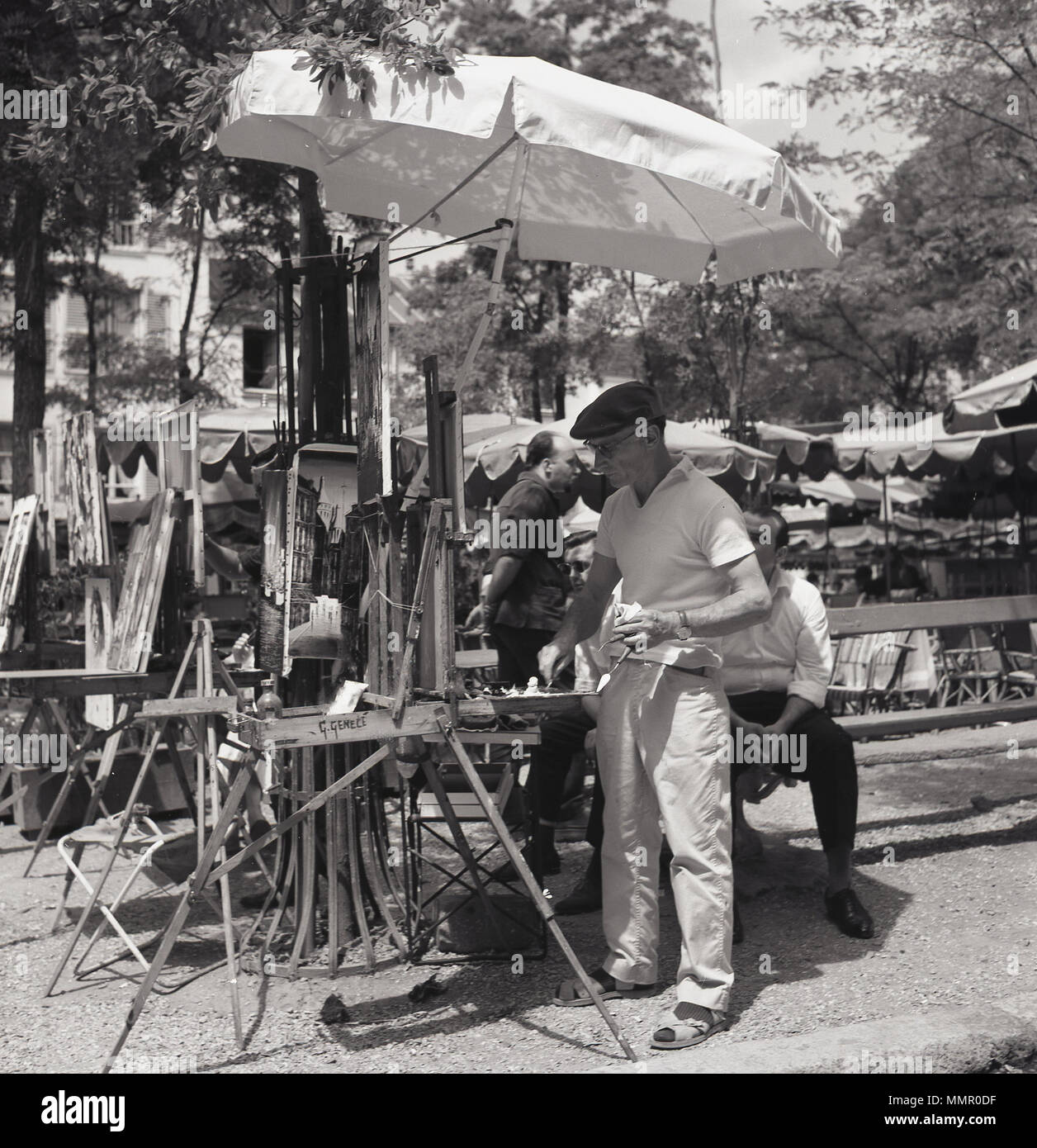 Durante el verano de 1950, en París, y de un artista de la calle bajo un paraguas trabajando en su caballete en la plaza arbolada Place du Tertre en Montmartre. La plaza principal en Montmartre, en la parte norte de la ciudad, es famosa por su historia artística y es un importante destino turístico en París. Foto de stock