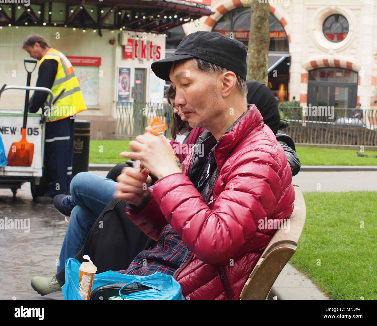 Un hombre sentado en un banco comiendo una máquina de paleta de helado en Leicester Square, Londres vistiendo una capa gruesa con un limpiador de la calle en el fondo Foto de stock