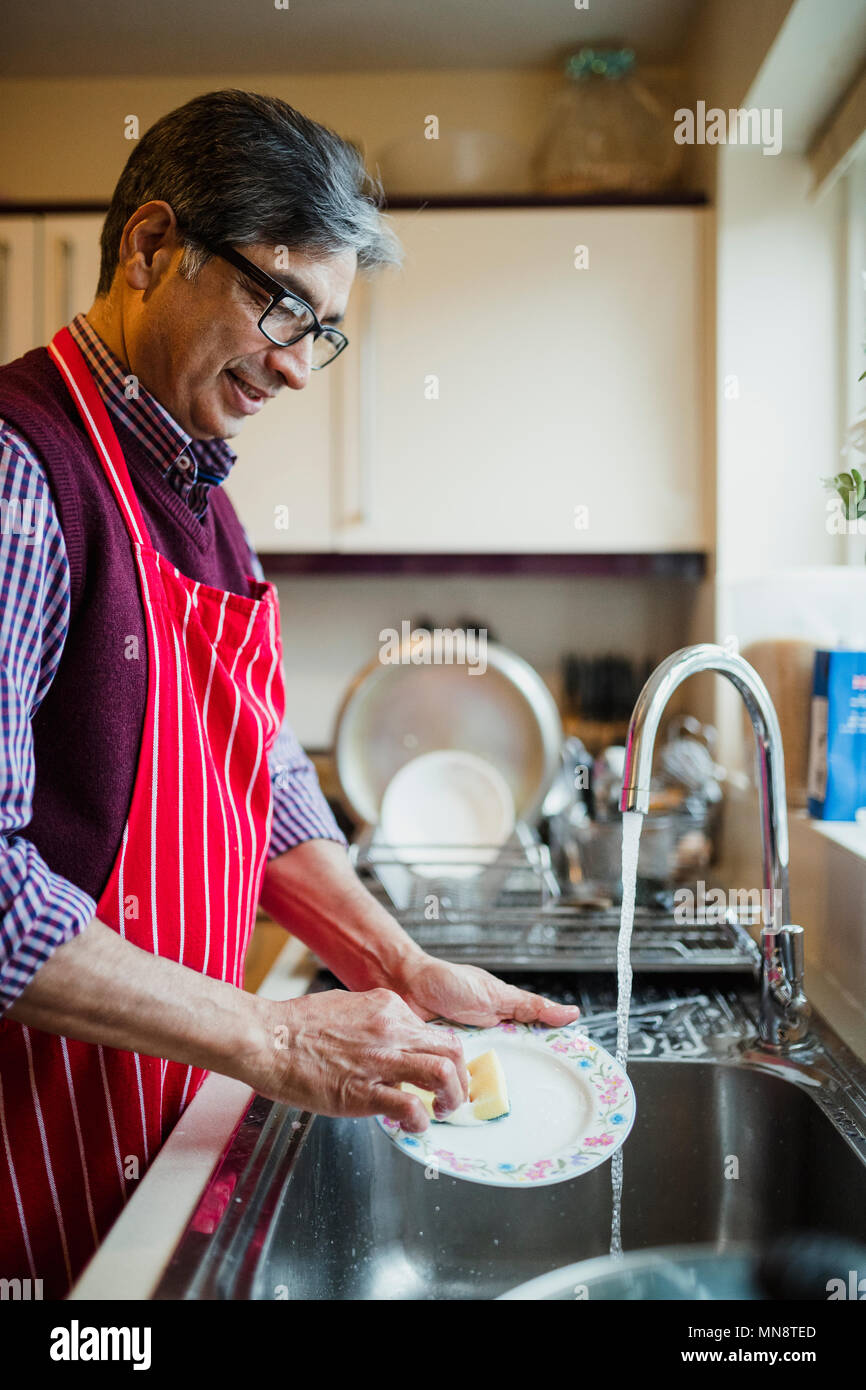 Hombre maduro está lavando los platos en la cocina de su casa. Él es el uso  de un delantal, mientras que el lavado de una pequeña placa en el fregadero  Fotografía de