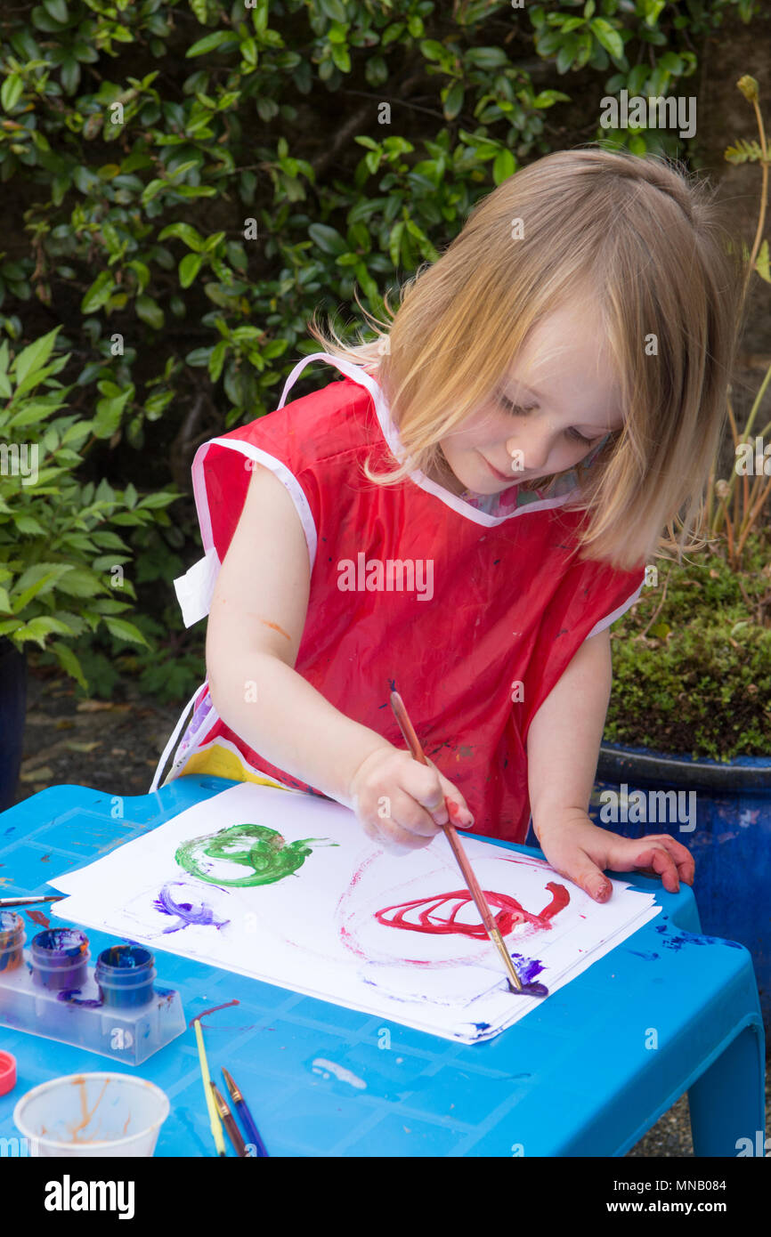 Dos años de edad, niña pintar imágenes y patrones con pinturas de acuarelas en jardín, conseguir pintura en las manos. Ropa protectora. Foto de stock