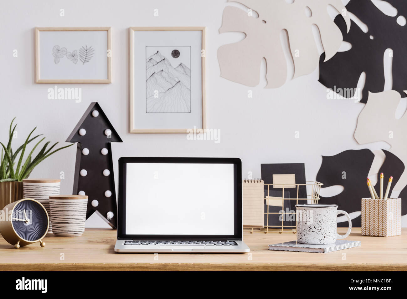 Portátil con pantalla blanca sobre un mostrador de madera con reloj, taza y lápices así como decoraciones de pared Foto de stock