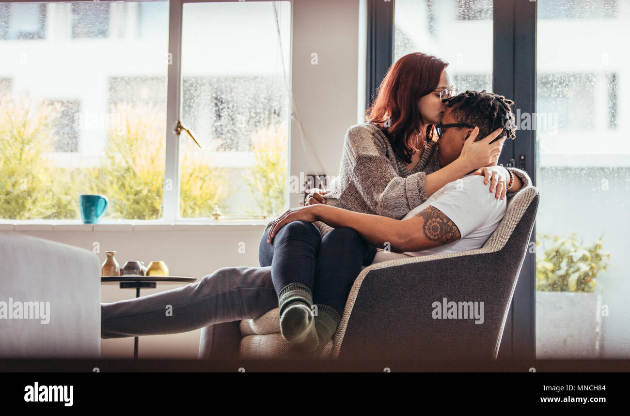 Mujer besar novio frente con amor mientras está sentado en una silla. Romántica pareja interracial en casa. Foto de stock