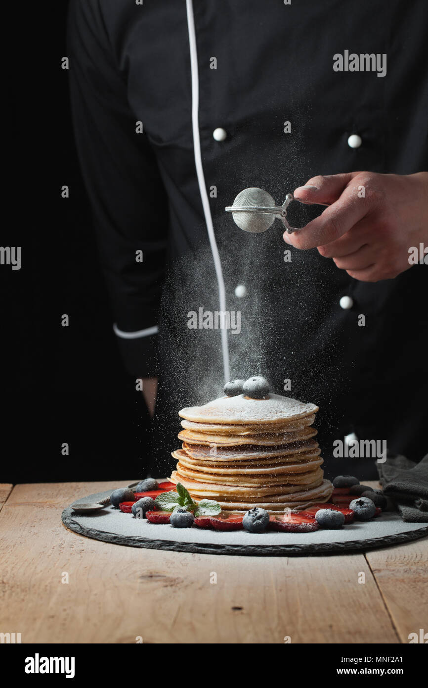 Sirve crepes con azúcar en polvo y bayas. Chef mujer mano. La comida aún hermosa vida. tonos ligeramente oscuro, imagen de fondo negro con área de texto. Vista horizontal Foto de stock