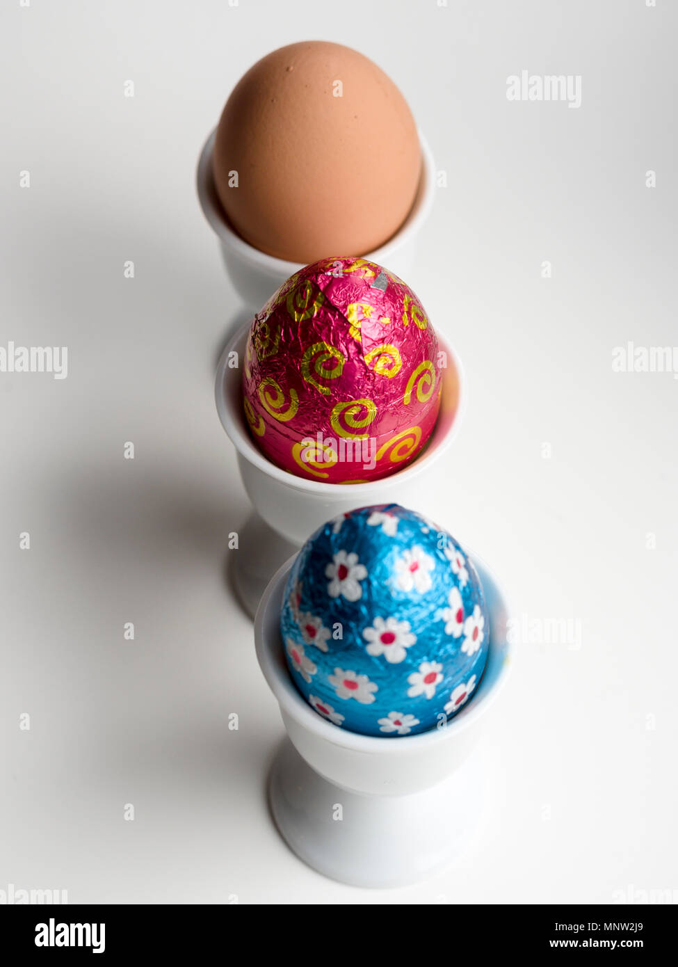 Huevos de Pascua de chocolate marrón y un huevo de gallina en el huevo copas: una fila de tres huevos en tres tazas de huevo de porcelana blanca sobre un fondo blanco. Foto de stock