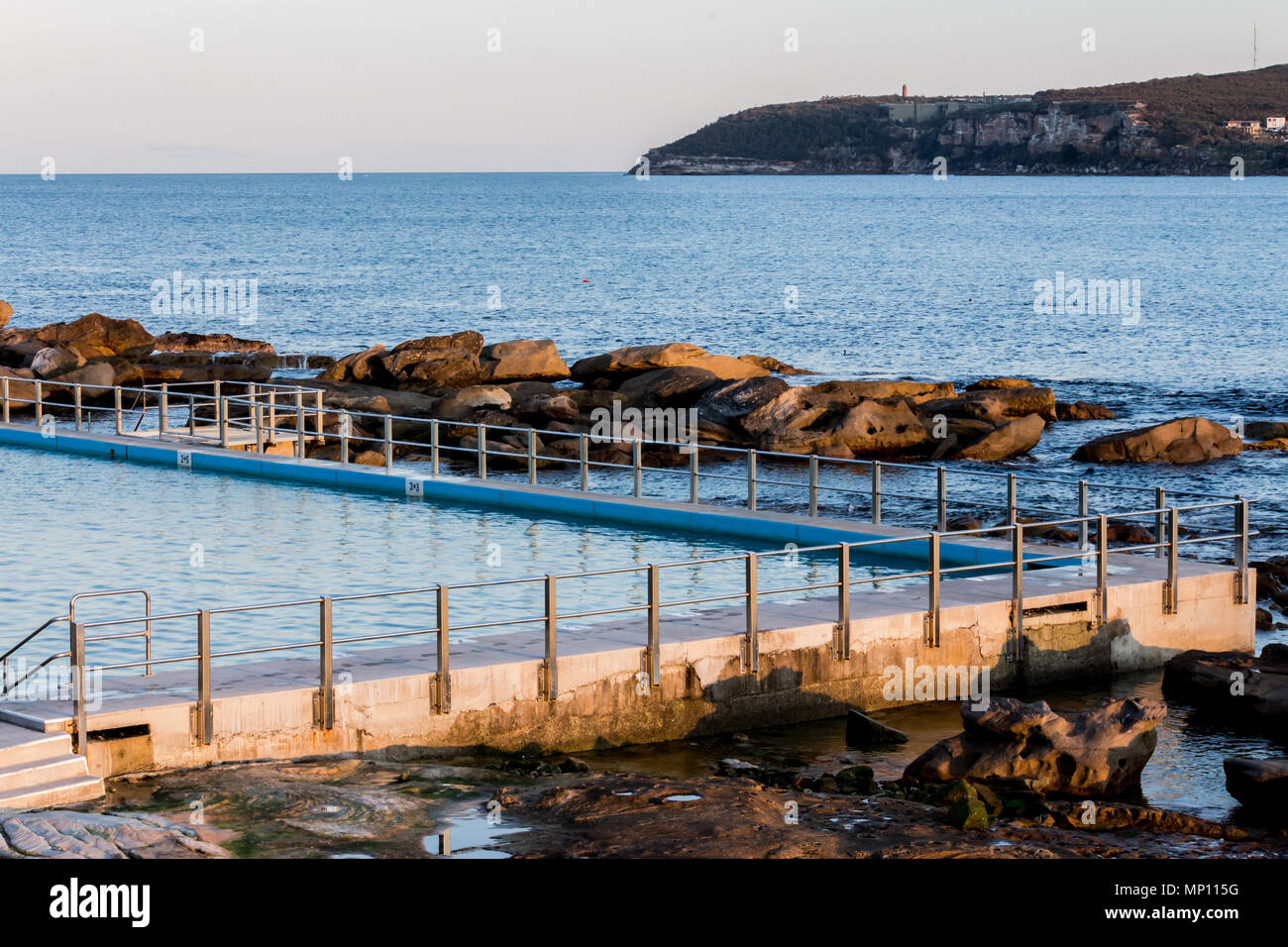 Piscina oceánica costera con la barandilla de metal set de esgrima contra el agua azul en calma y grandes rocas expuestas Foto de stock