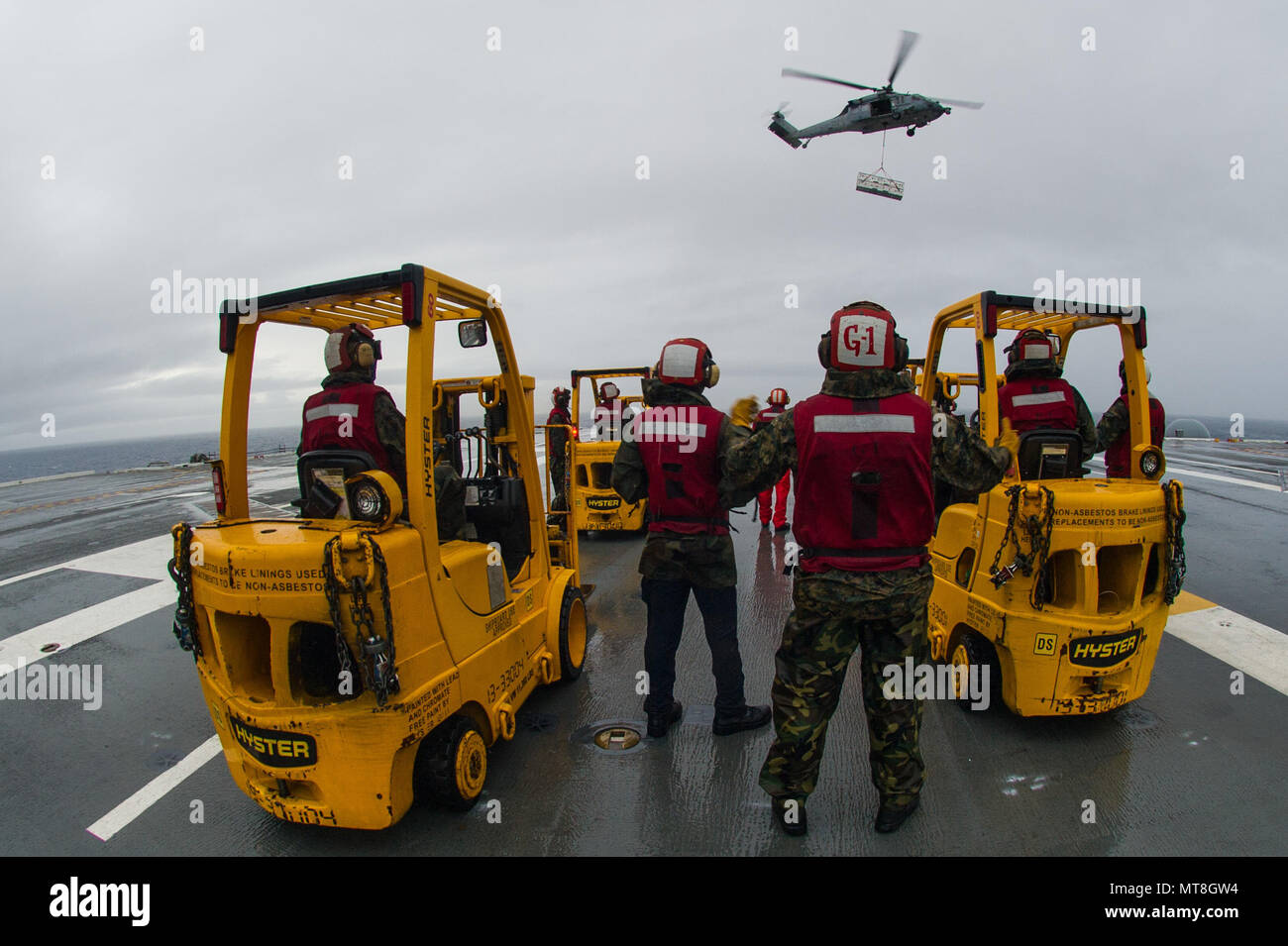 180514-N-OY799-0103 las aguas al sur de Japón (14 Mayo 2018) departamento de armas marineros de permanecer como un MH-60S Sea Hawk, asignado al comando de Transporte Marítimo Militar (MSC) carga seca/municiones buque USNS César Chávez (T-AKE 14), ofrece detonar en la cubierta de vuelo a bordo de la Marina desplegadas, el portaviones USS Ronald Reagan (CVN 76), como parte de una reposición en alta mar durante las pruebas de mar. Los civiles no combatientes, con tripulación de barco, operado por la MSC, proporciona el combustible, alimentos, municiones, piezas de repuesto, el correo y otros suministros a los buques de la Marina en todo el mundo. Ronald Reagan, el buque insignia de transportista Stri Foto de stock