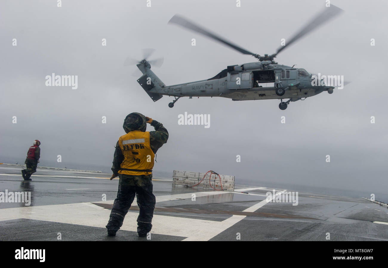 180514-N-OY799-0218 las aguas al sur de Japón (14 de mayo de 2018) la aviación Boatswain's Mate (manejo) de primera clase, Thinh Dai saluda a un MH-60S Sea Hawk, asignado al comando de Transporte Marítimo Militar (MSC) carga seca/municiones buque USNS César Chávez (T-AKE 14), después de lanzar las municiones en la cubierta de vuelo a bordo de la Marina desplegadas, el portaviones USS Ronald Reagan (CVN 76), como parte de una reposición en alta mar durante las pruebas de mar. Los civiles no combatientes, con tripulación de barco, operado por la MSC, proporciona el combustible, alimentos, municiones, piezas de repuesto, el correo y otros suministros a los buques de la Marina en todo el mundo. Ronald Reagan. Foto de stock