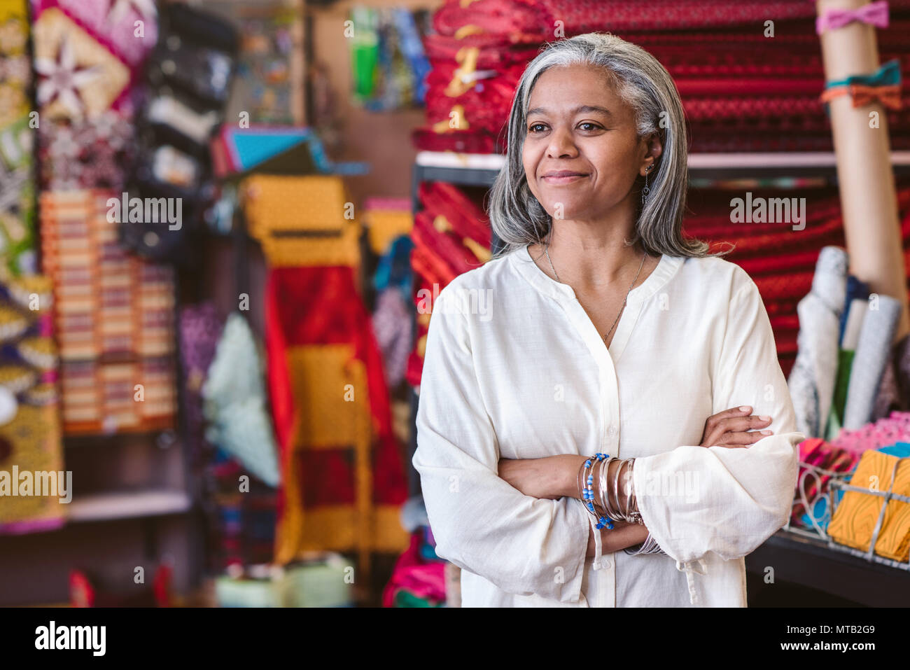 Sonriente tejido maduro propietario de una tienda de pie con los brazos cruzados rodeado de estanterías y de coloridas telas y textiles Foto de stock
