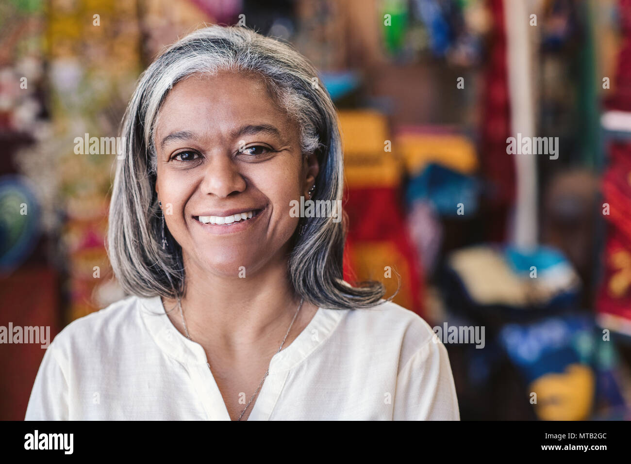 Retrato de un sonriente maduro propietario de una tienda de tela de pie en su tienda con coloridas telas y textiles en estanterías en el fondo Foto de stock
