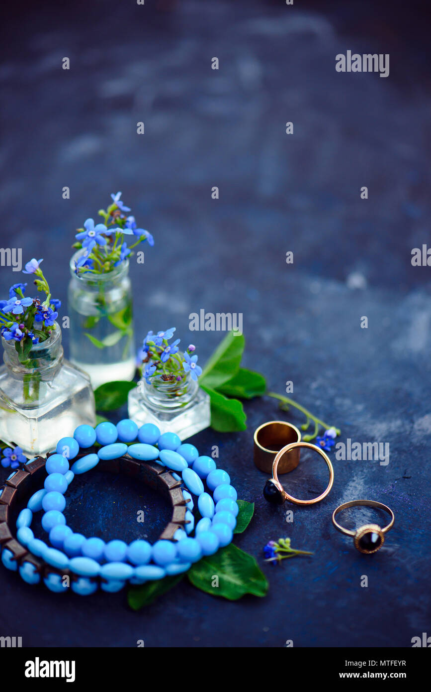 Blue Bead pulseras y anillos de oro en un estilo femenino concepto con flores azules en pequeñas botellas sobre un fondo oscuro. Accesorios de verano hermosa con espacio de copia Foto de stock