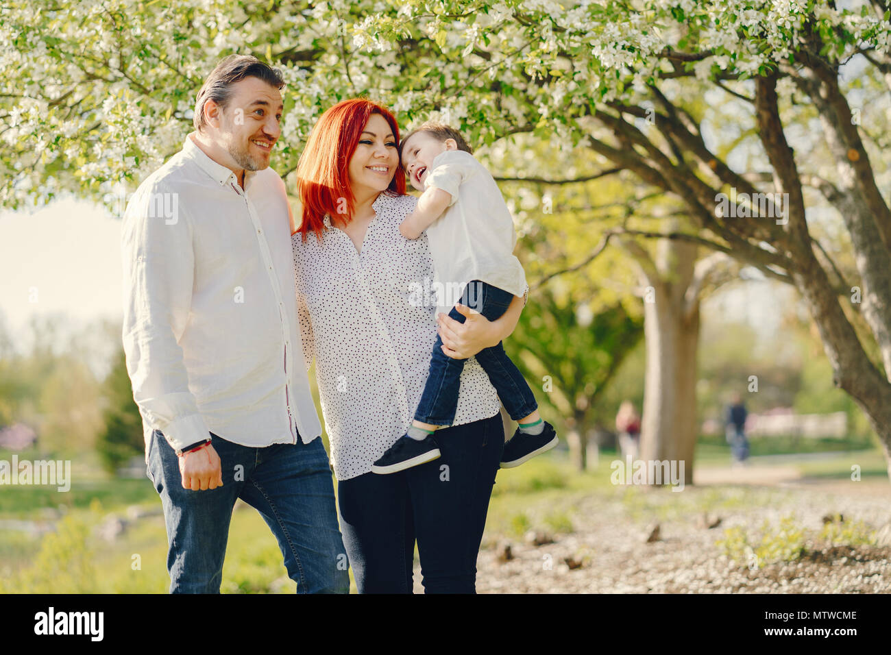 Hermoso y elegante redhead mom en una blusa blanca de pie con su bonito  hombre vestido con una camisa blanca y pantalones de mezclilla juegan con  sus littl Fotografía de stock -