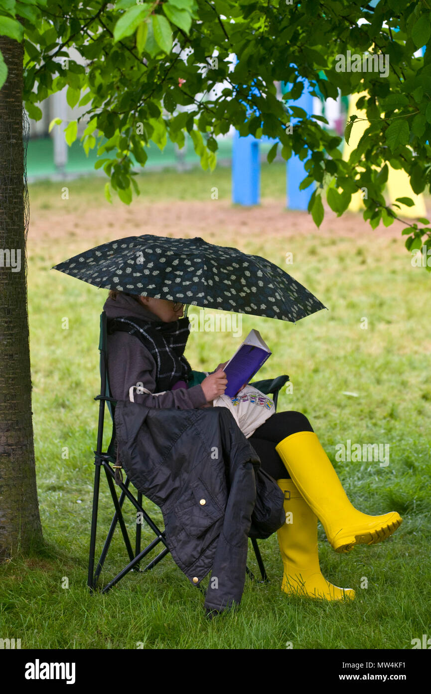 Mujer leyendo un libro con sombrilla y vistiendo amarillo wellies sentado debajo de un árbol en el jardín en 2018 FESTIVAL DE HENO HENO-on-Wye Gales Powys UK Foto de stock