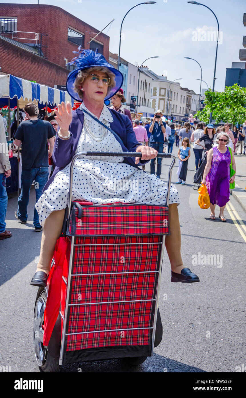 Un hombre vestido como una mujer vieja montando un carrito de la compra es en realidad un ejecutante de la calle entretenido multitud en un mercado de primavera en Hammersmith & Fulham Foto de stock