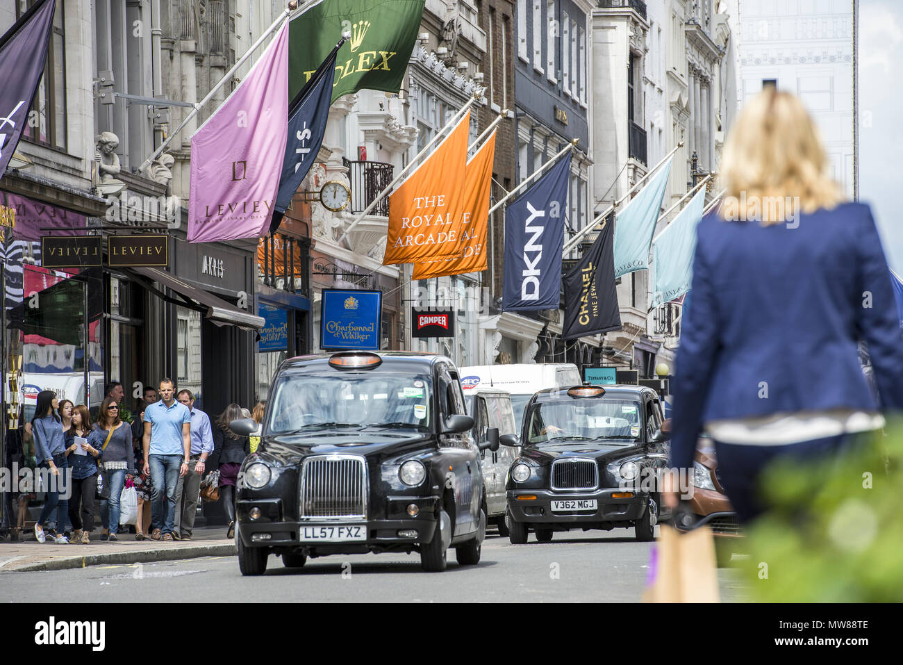 Un día ajetreado en el distrito comercial de Mayfair. Una mujer con bolsas de compras camina mientras los taxis negros conducen por la calle bordeada de banderas de Marca Foto de stock