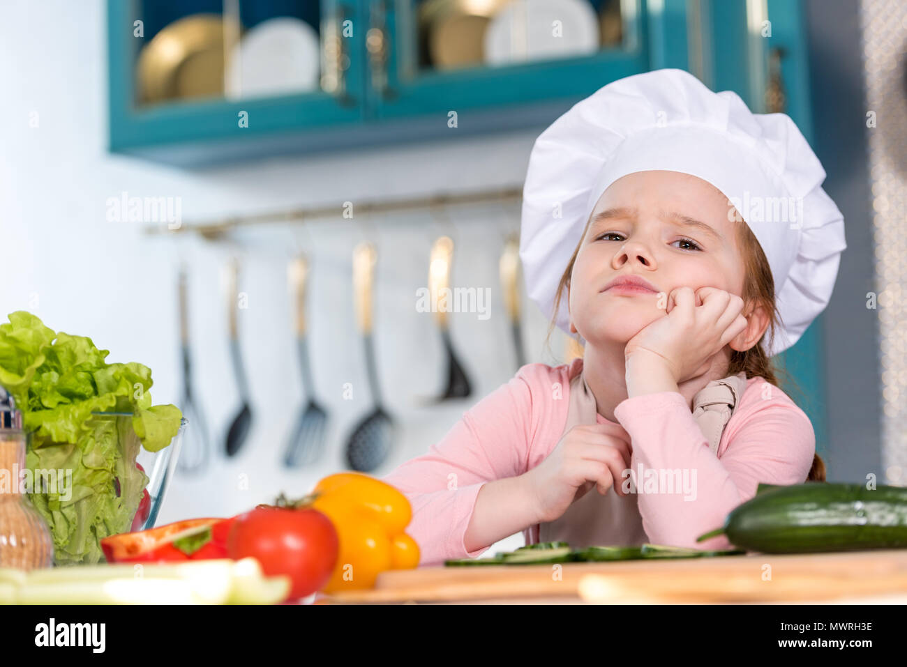 Niño aburrido en el chef hat sentado con la mano en el mentón en la cocina Foto de stock