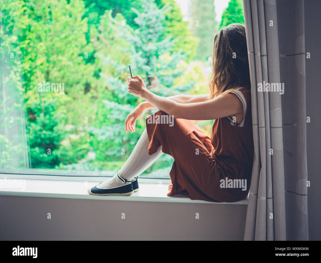 Un joven está sentado al lado de la ventana mirando los árboles fuera mientras utiliza su teléfono inteligente. Foto de stock