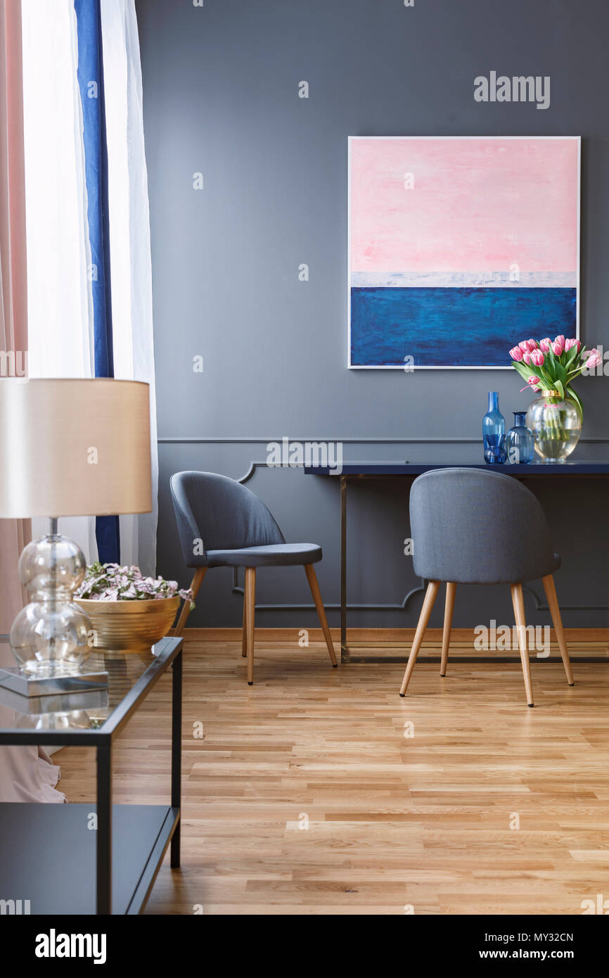 Foto real de un amplio comedor interior con una pintura azul y rosa sobre  una pared gris, encima de una mesa y dos sillas y una mesa con lámpara  Fotografía de stock -