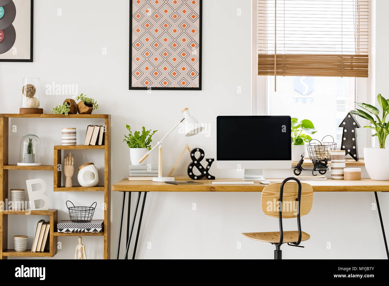 Foto real de un escritorio con una pantalla de ordenador, lámpara y ornamentos de pie con una silla junto a un rack en una sala de trabajo con carteles en una pared y ventana Foto de stock