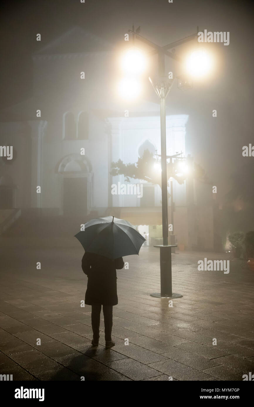 Persona con sombrilla pararse bajo la lámpara de la calle en frente de la catedral por la lluvia y la niebla en la noche, la costa de Amalfi, Ravello, Campania, Italia, Europa Foto de stock