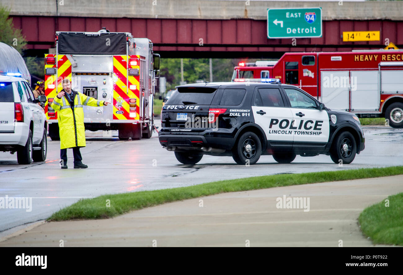 El 18 de mayo de 2018 Stevensville MI EE.UU.; un hombre de la policía en ropa de lluvia dirige el tráfico en la escena de un accidente grave, con camiones de bomberos esperando detrás de él Foto de stock