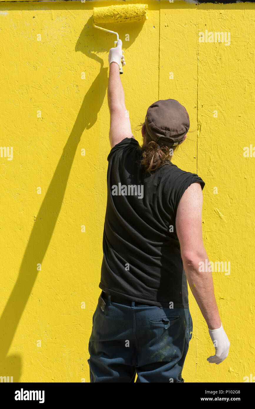 Un hombre vestido con ropa negra y una gorra de béisbol con un rodillo para pintar el lateral de un edificio o pared en una pintura de color amarillo pintar