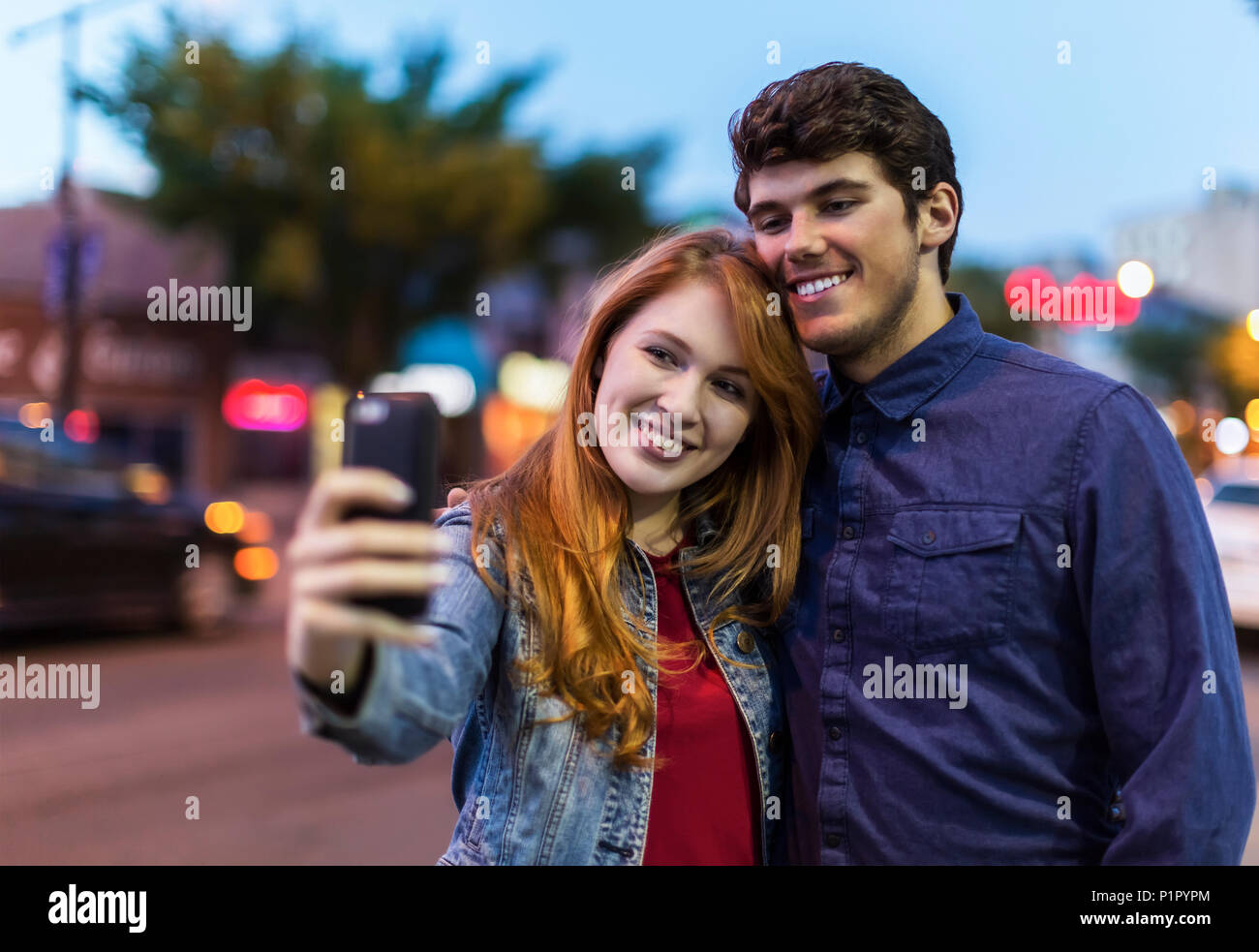 Una joven pareja están junto a una calle de la ciudad al atardecer tomando un auto-retrato con un teléfono inteligente, Edmonton, Alberta, Canadá Foto de stock