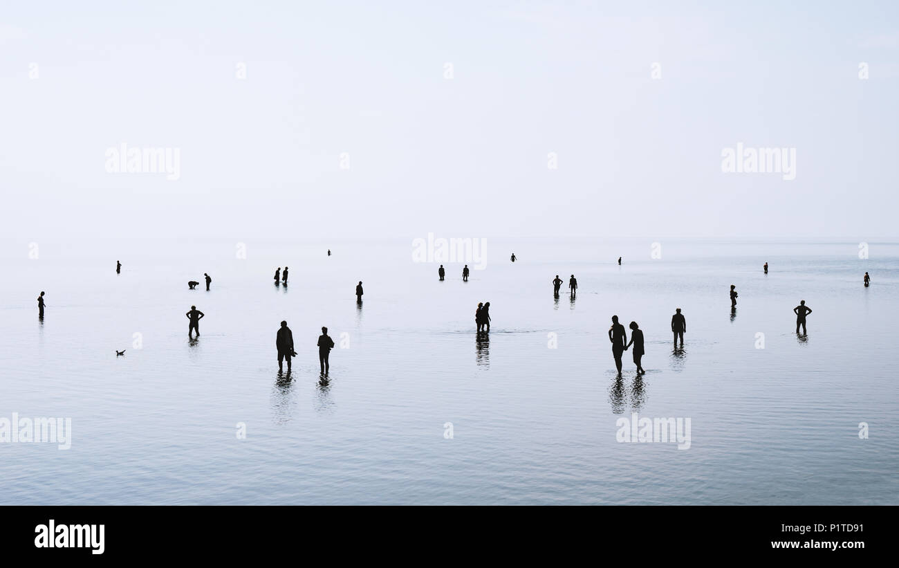 Grandes grupos de gente o multitud de pie caminar y nadar en aguas superficiales en la costa del mar del norte de Alemania durante el reflujo, siluetas retroiluminado Foto de stock