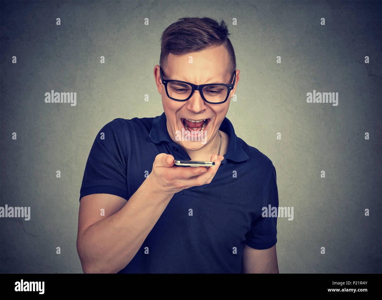 Joven Hombre frustrado en anteojos gritando smartphone funciona mal sobre fondo gris. Foto de stock