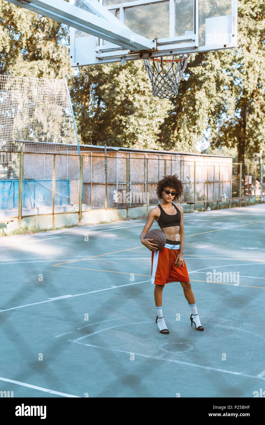 Joven Mujer afroamericana en vestimenta deportiva y tacones altos  sosteniendo una pelota de baloncesto en el campo de deportes Fotografía de  stock - Alamy