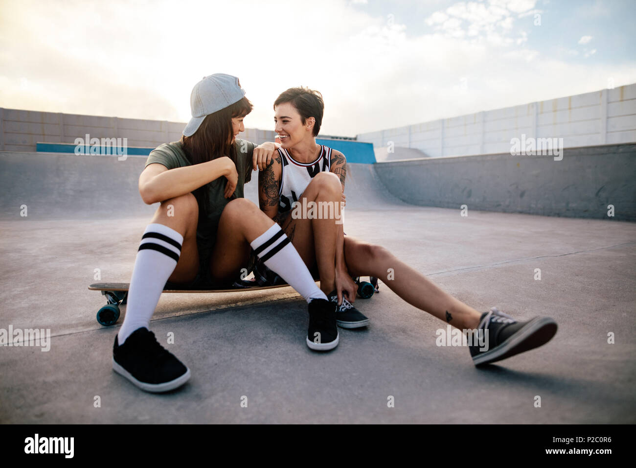 Dos amigas disfrutando de pasar tiempo juntos en el parque de skate. Mujer skateboarders sentado en placa larga y mirando el uno al otro. Foto de stock