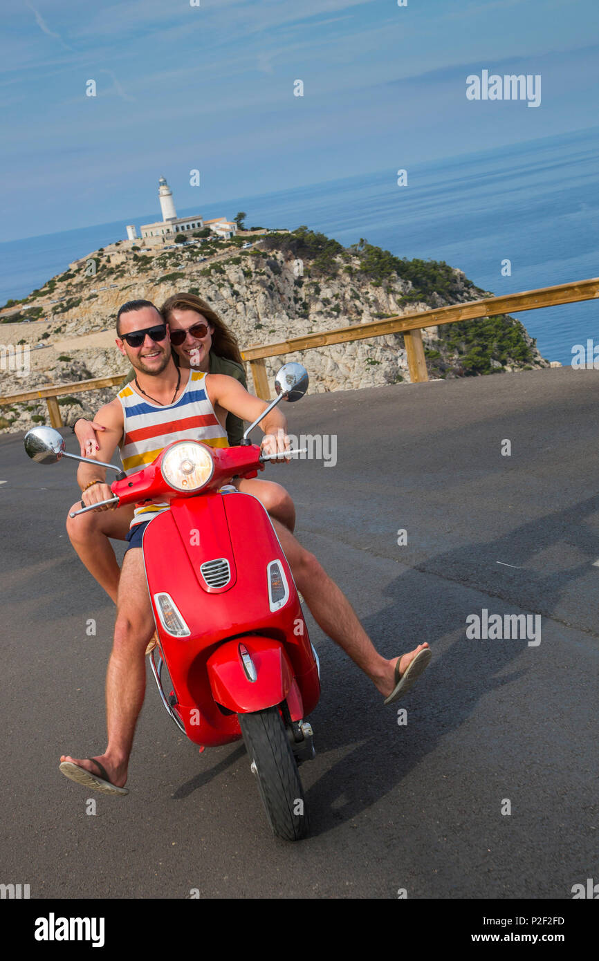 Pareja joven montando una Vespa scooter rojo en la carretera a lo largo de la península de Cap de Formentor con el Faro de Formentor, Palma detrás del faro Foto de stock