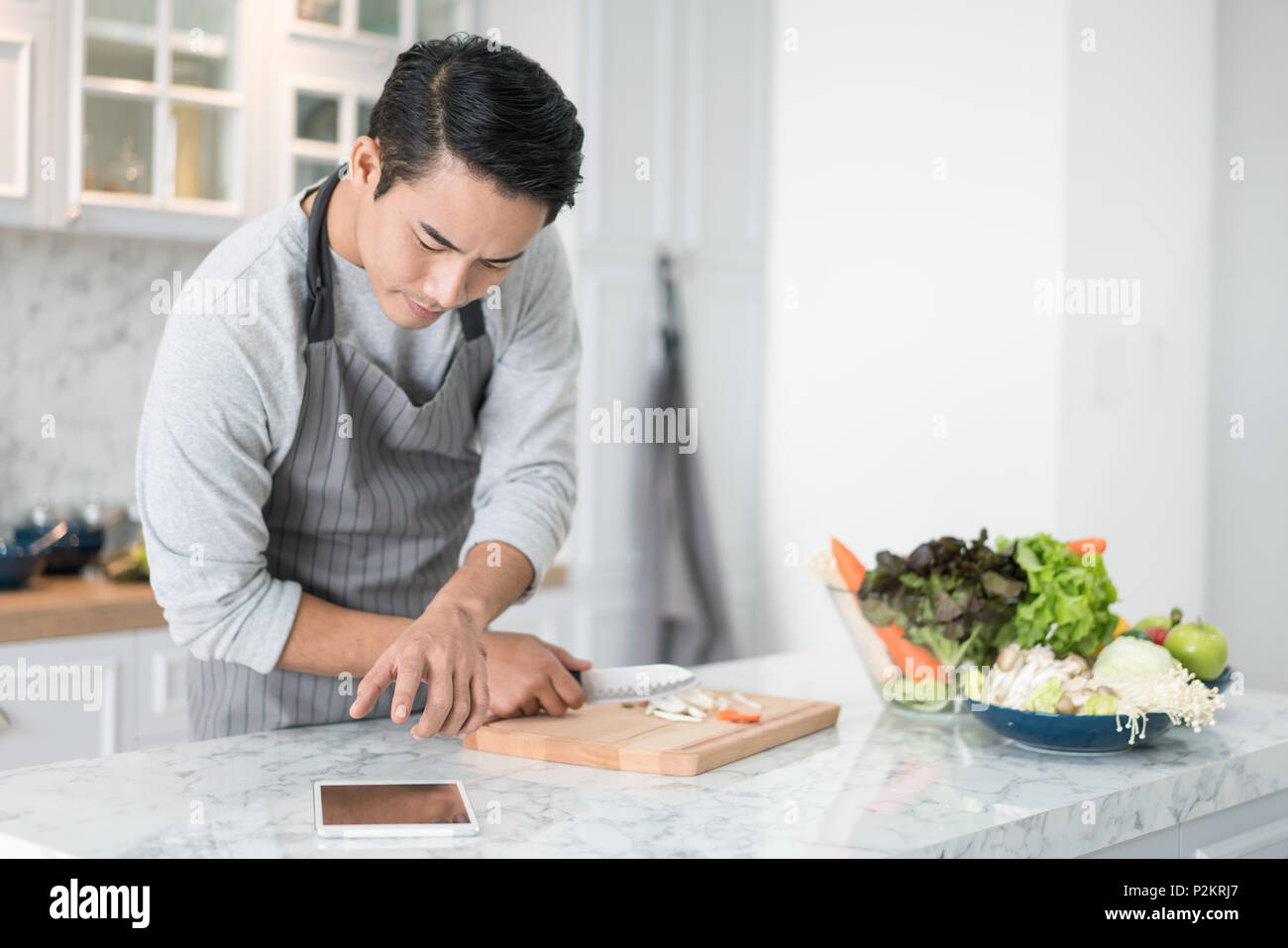 Asian confundido hombre leyendo su tableta con una mirada pensativa pensativa mientras está de pie en la cocina de su casa mientras cocina y preparar una comida de una variedad o Foto de stock