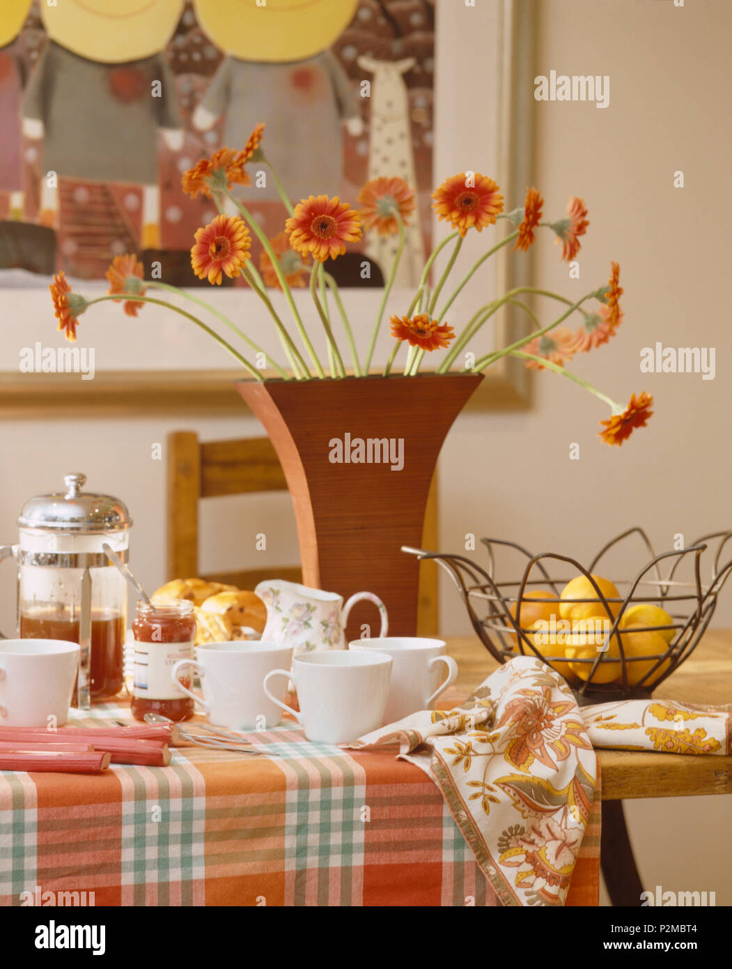 Close-up de jarrón de gerberas naranjas de mesa de comedor con marcada paño y cafetera junto a blanco tazas Foto de stock