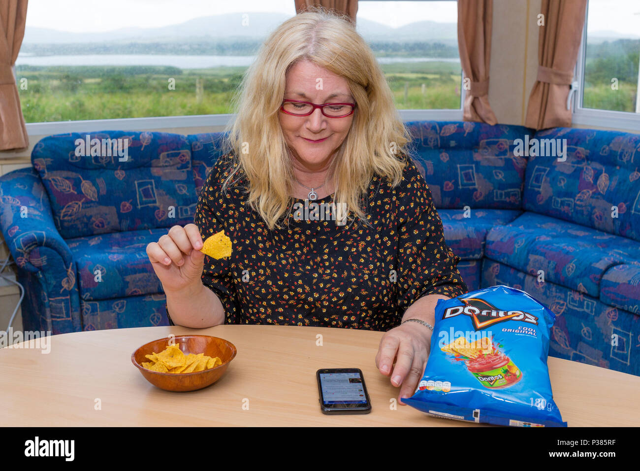 Mujer rubia comiendo Doritos desde una bolsa de tamaño compartido y un pequeño cuenco Foto de stock