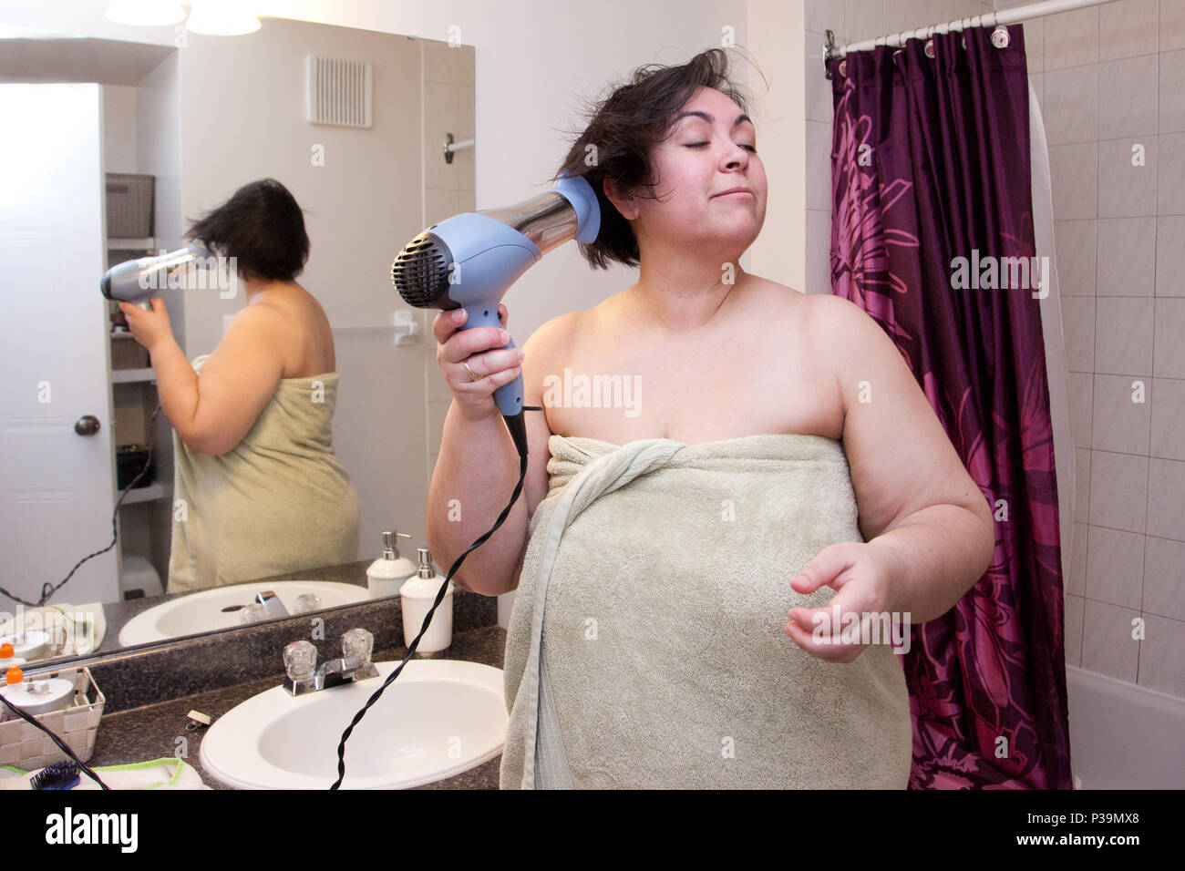 El secado de cabello envuelto en una toalla, una mujer permanece en su cuarto de baño, preparándose para salir Foto de stock