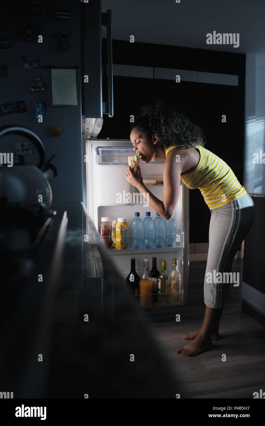 Afroamericana haciendo midnight snack en el hogar. Ella come un bocadillo y busca los alimentos en el refrigerador durante la noche. Foto de stock