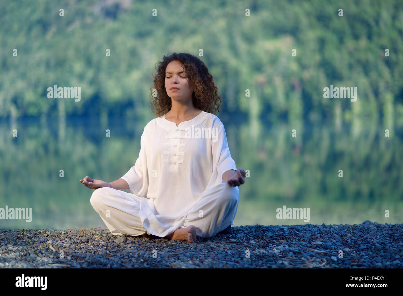 Mujer joven en traje de seda blanco meditando en el amanecer por el lago en la naturaleza Foto de stock