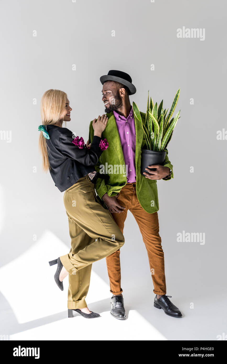 Sonriente pareja interracial elegante con el verde de las plantas en maceta posando sobre fondo gris Foto de stock