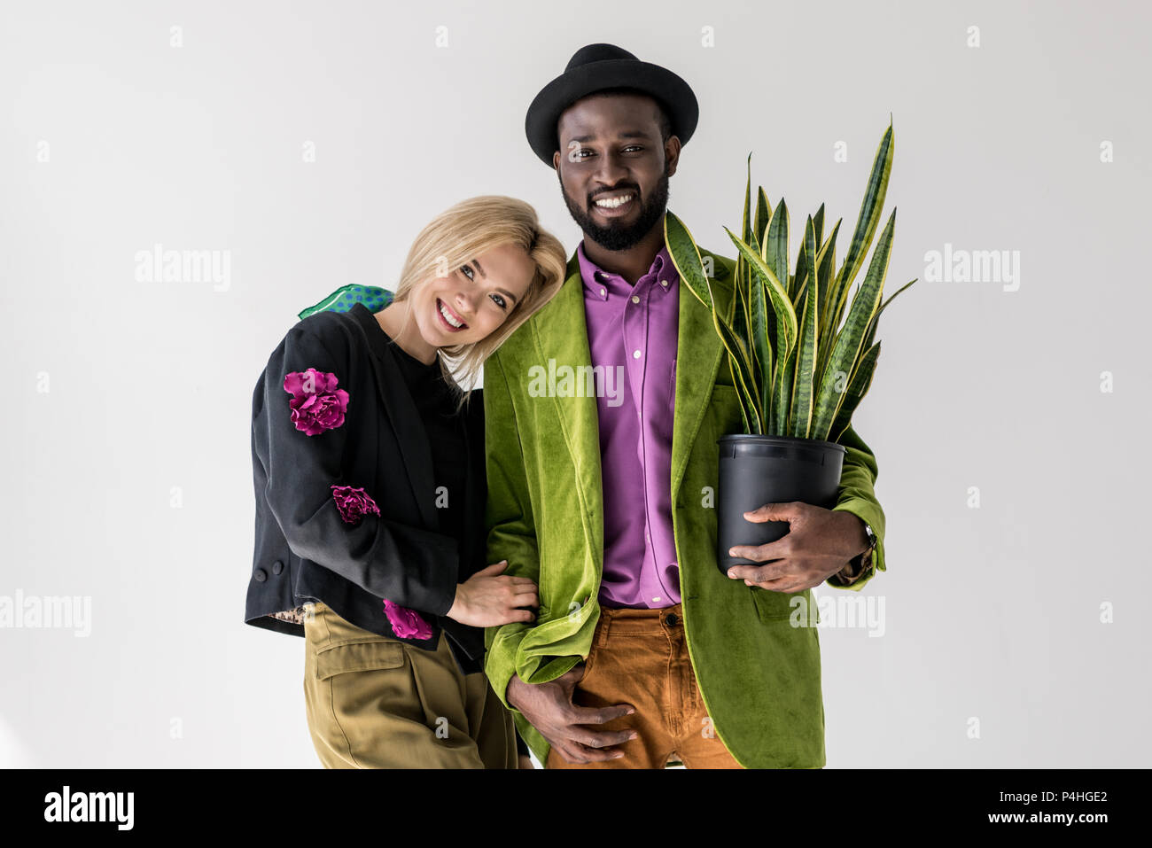 Retrato de sonriente pareja interracial elegante con el verde de las plantas en maceta posando aislados en gris Foto de stock